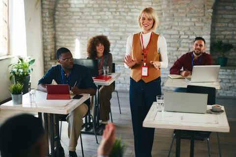 A polished female presenter engages a diverse team in a bright coworking space. Attendees listen, take notes, and interact with laptops