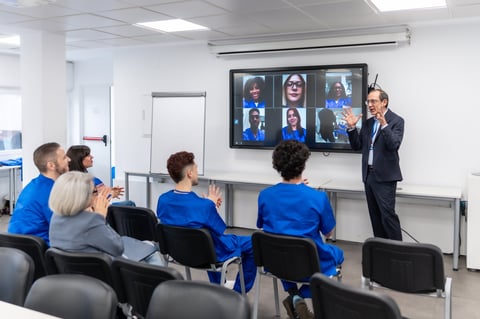 Professor lecturing to a group of students in a classroom, with other students joining remotely through a video conference on a large screen