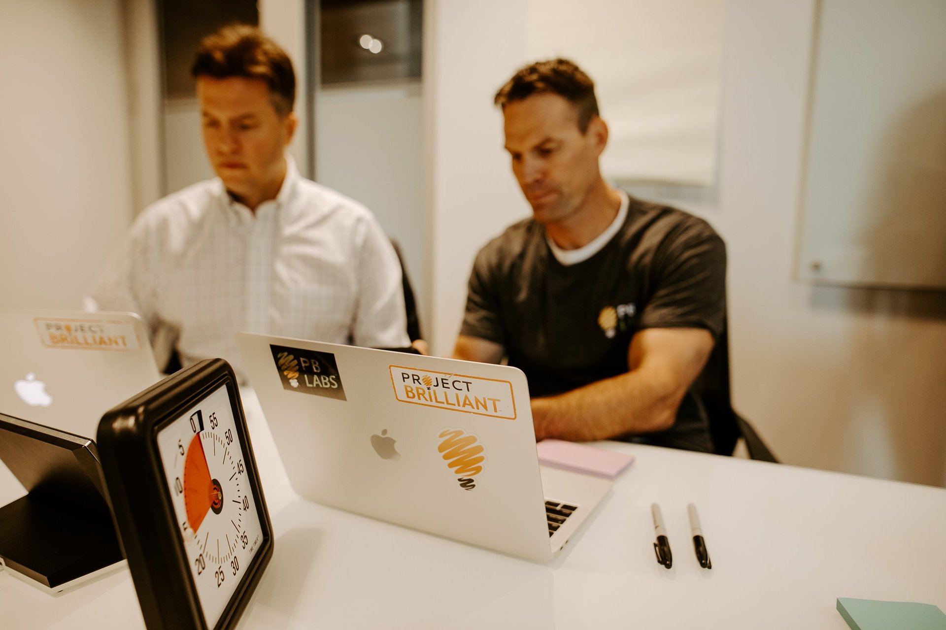 Two men working at a table with laptops and tech equipment in an office environment