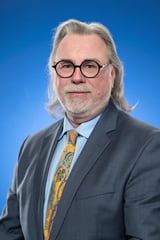 Professional headshot of a middle-aged man with long gray hair, glasses, and a goatee wearing a charcoal suit and gold patterned tie against a blue background.