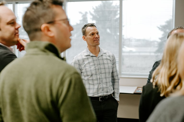 Man in plaid shirt standing in office with group of people facing him by window with city skyline view
