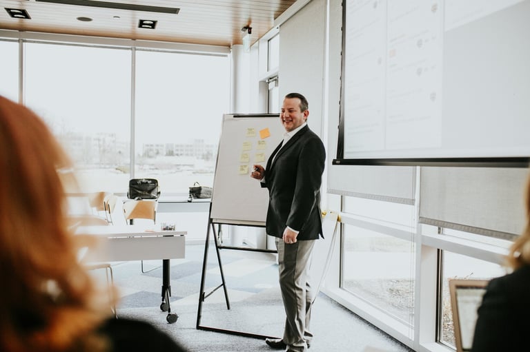 Businessman in dark suit presenting at a flipchart during a meeting in a modern office with large windows.