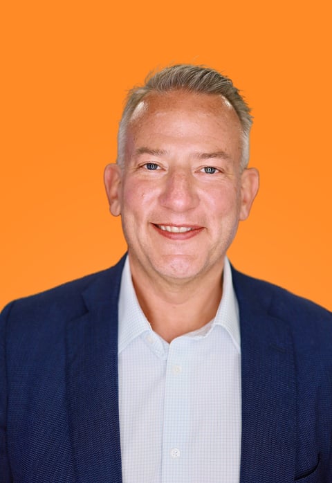 Professional headshot of a smiling middle-aged man wearing a dark blue blazer and light blue shirt against an orange background